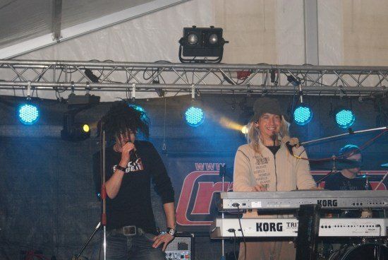 Zwei Musiker treten auf der Bühne unter blauen Lichtern auf, einer spielt ein Keyboard und der andere singt in ein Mikrofon.