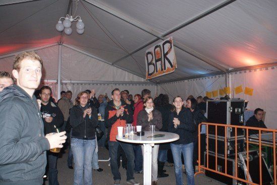 Eine Gruppe von Menschen steht in einem Zelt, einige klatschen, mit einem Bar-Schild darüber und einem Tisch mit Gläsern davor.