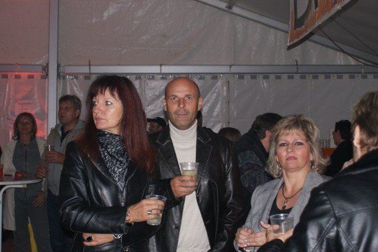 Eine Gruppe von Menschen in einem Zelt, zwei Frauen und zwei Männer, alle mit Getränken in der Hand, mit einem Mann in einem weißen Rollkragen in der Mitte.