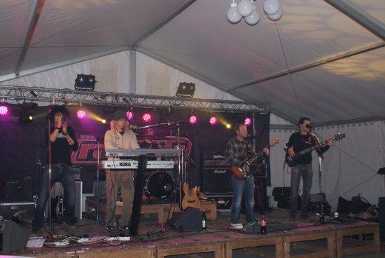 Eine Band spielt auf einer Bühne unter einem Zelt mit einem Schlagzeug, einer Tastatur und Gitarren. Die Bühne ist mit roten und lila Lichtern beleuchtet.