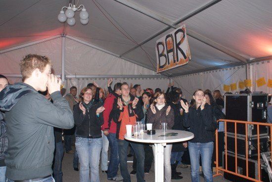 Eine Gruppe von Menschen versammelt sich in einem Zelt, einige klatschen und andere trinken. Ein Banner mit der Aufschrift 'BAR' hängt von der Decke.