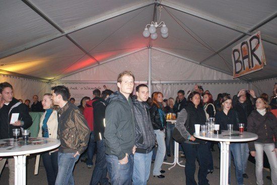 Eine Gruppe von Menschen steht in einem Zelt mit einem Bar-Schild. Einige halten Getränke und andere führen Gespräche. Das Zelt ist gut beleuchtet mit Deckenleuchten.