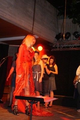 Eine Frau in einem roten Kleid steht auf der Bühne mit einem Klavier, während andere Frauen in Kleidern und Hüten hinter ihr stehen. Scheinwerfer und ein Mikrofon sind sichtbar.
