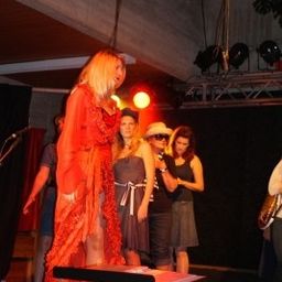 Eine Frau in einem roten Kleid steht auf der Bühne mit einem Klavier, während andere Frauen in Kleidern und Hüten hinter ihr stehen. Scheinwerfer und ein Mikrofon sind sichtbar.