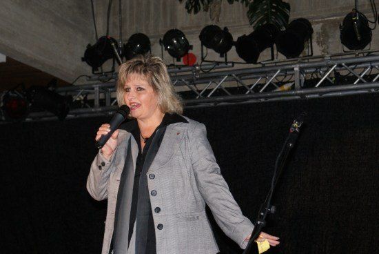 Eine Frau in einem grauen Blazer und einem schwarzen Shirt singt auf der Bühne und hält ein Mikrofon. Scheinwerfer sind an einem Metallrahmen über ihr montiert.