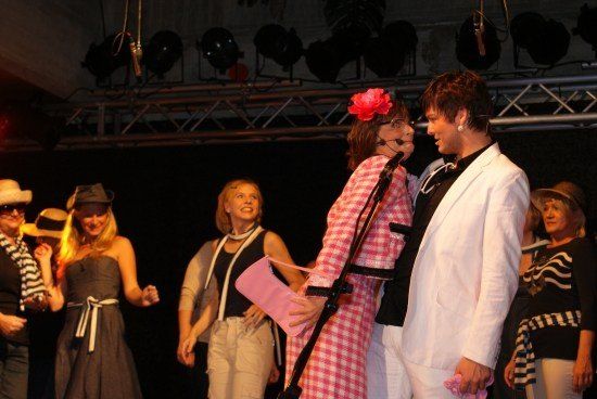 Eine Gruppe von Menschen auf der Bühne, eine Frau in einem rosa karierten Kleid hält ein Mikrofon, eine andere Frau in einem schwarzen Kleid und ein Mann in einem weißen Anzug.