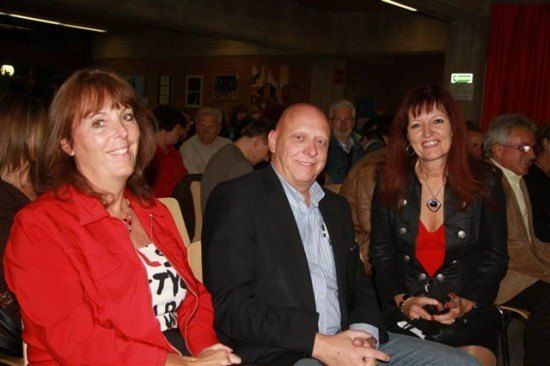 Drei Menschen sitzen auf Stühlen in einem Raum und lächeln für ein Foto. Ein Mann in einem Anzug sitzt zwischen zwei Frauen. Hinter ihnen sitzen mehrere Menschen, und ein roter Vorhang ist im Hintergrund.