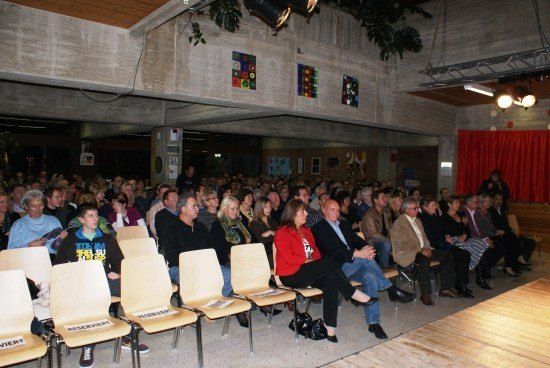 Ein großes Publikum sitzt in Reihen in einem Raum mit Bühne, rotem Vorhang und dekorativer Wandkunst.