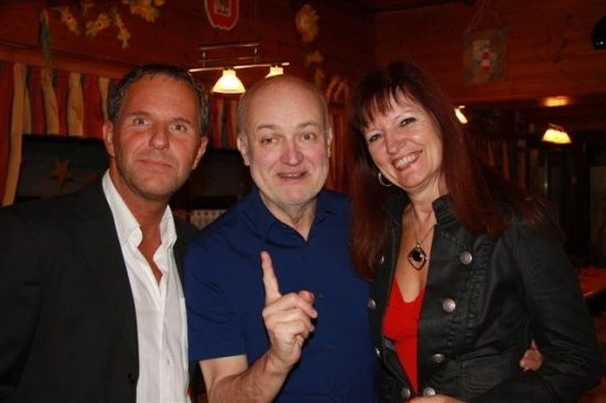 Drei Menschen lächeln und posieren für ein Foto in einem Restaurant. Der Mann in der Mitte zeigt mit dem Finger nach oben.