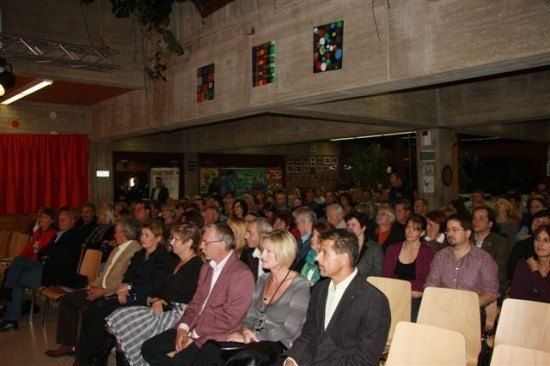 Eine große Gruppe von Menschen sitzt in Stühlen in einem Raum mit Bühne und farbenfrohen Gemälden an der Wand.