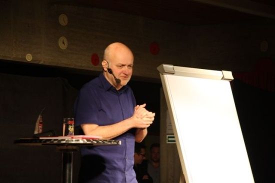 Ein Mann in einem blauen Hemd steht hinter einem Podium und spricht in ein Mikrofon. Ein Whiteboard befindet sich hinter ihm.