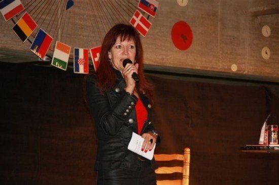 Eine Frau in einer schwarzen Lederjacke spricht in ein Mikrofon vor einer Wand mit Flaggen und einem roten Kreis.