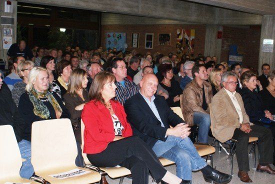 Eine große Gruppe von Menschen sitzt in Stühlen in einem Raum, möglicherweise bei einer Besprechung oder Präsentation. Einige tragen Brillen, und einige lächeln. Der Raum hat Ziegelwände und eine Decke mit Lichtern.