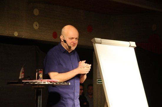 Ein kahlköpfiger Mann in einem blauen Hemd und einem Mikrofon hält eine Präsentation, während er vor einer Tafel steht.