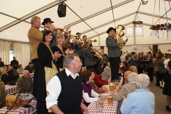 Ein Blasorchester spielt in einem Zelt, während eine Menschenmenge an Tischen sitzt, einige klatschen und andere Bier trinken. Das Zelt hat weiße Wände und ein Metallgerüst.