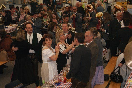 Eine große Gruppe von Menschen in einem Saal, einige spielen Musikinstrumente, andere unterhalten sich, mit Tischen und Bänken im Hintergrund.
