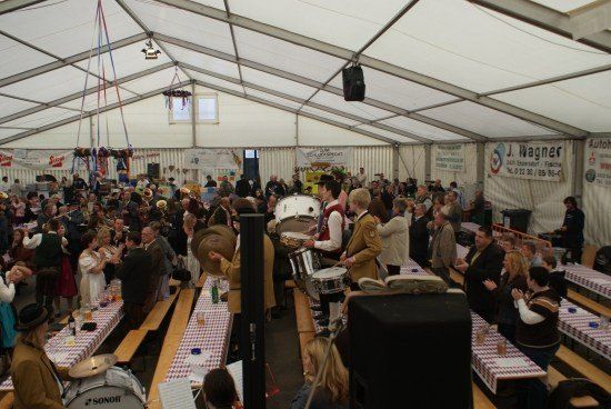 Eine große Indoor-Versammlung mit Menschen, die um Tische herum sitzen, einige Schlagzeug spielen und andere klatschen. Die Umgebung ist ein Zelt mit Bannern und Lautsprechern.