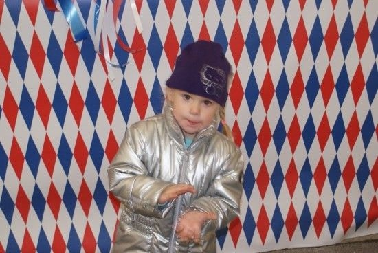 Ein kleines Mädchen in einer silbernen Jacke und einer lila Mütze steht vor einem roten, weißen und blauen Rautenmuster-Hintergrund.