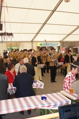 Eine Blaskapelle spielt in einem Zeltbereich mit Tischen und Stühlen, wo Menschen sitzen und zuschauen. Das Zelt ist mit rot-weiß-blau karierten Tischdecken dekoriert.