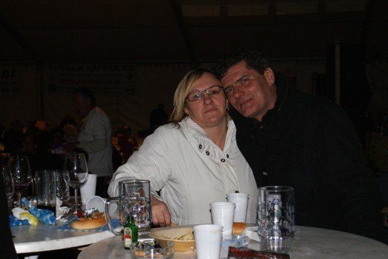 Ein Paar sitzt an einem Tisch mit Essen und Getränken und lächelt für ein Foto in einem schwach beleuchteten Raum.