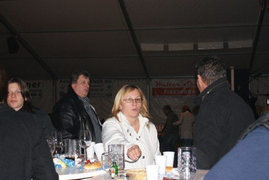 Eine Gruppe von Menschen sitzt in einem Zelt um einen Tisch herum, wahrscheinlich bei einer Veranstaltung. Die Frau in der Mitte schaut auf etwas auf dem Tisch. Es gibt Gläser, Tassen und Essen auf dem Tisch. Im Hintergrund stehen auch Menschen.