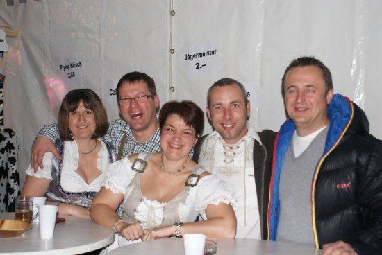 Fünf Menschen lächeln und posieren für ein Foto in einem Zelt mit weißem Hintergrund. Zwei Frauen tragen traditionelle deutsche Kleidung. Ein Mann auf der rechten Seite trägt eine schwarze Jacke mit blau-gelbem Streifen. Es gibt Tassen auf dem Tisch vor ihnen.