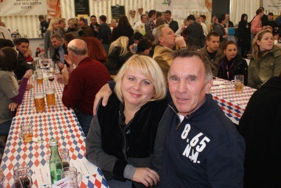 Ein Mann und eine Frau sitzen an einem Tisch mit Biergläsern und lächeln für ein Foto, mit einer großen Menschenmenge im Hintergrund.