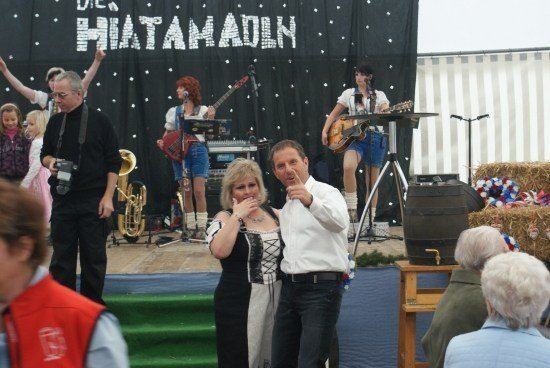 Eine Band spielt auf der Bühne mit einem Banner, das 'Die Hiatamadln' besagt. Ein Mann und eine Frau tanzen vor der Bühne, während eine Menge zuschaut.