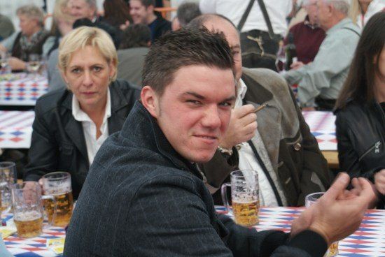 Ein Mann in einer schwarzen Jacke sitzt an einem Tisch mit einem Bierkrug, umgeben von anderen Menschen in einer festlichen Outdoor-Umgebung.