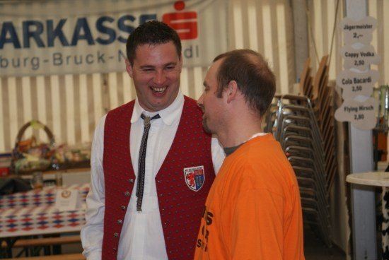 Zwei Männer stehen lächelnd voreinander vor einem Banner, das 'Markkass' steht. Der Mann auf der linken Seite trägt eine rote Weste mit Logo. Dahinter steht ein Tisch mit einem Korb und Stühlen.
