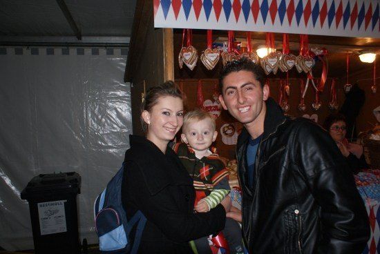 Eine dreiköpfige Familie, bestehend aus einem lächelnden Mann, einer Frau mit einem Kind und einem Baby, steht vor einem dekorierten Stand.