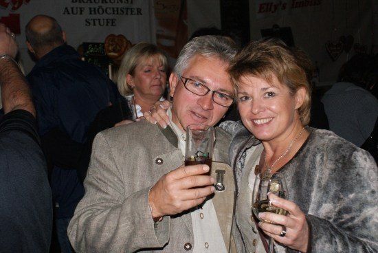 Ein Mann und eine Frau lächeln und halten Getränke in einer sozialen Umgebung, mit anderen Leuten im Hintergrund.