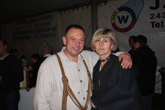 Ein Mann und eine Frau stehen lächelnd für ein Foto bei einer Veranstaltung. Der Mann trägt eine Hose mit Aufhängung und ein weißes Hemd, während die Frau einen schwarzen Pullover trägt. Hinter ihnen befindet sich ein Banner mit einem Logo und Kontaktinformationen.