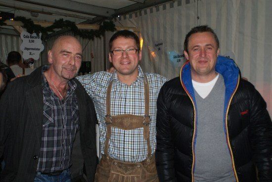 Drei Männer stehen zusammen in einer festlichen Umgebung, einer trägt traditionelle bayerische Kleidung mit Hosenträgern und einem karierten Hemd, ein anderer eine schwarze Jacke und der dritte eine schwarze Daunenjacke. Hinter ihnen befinden sich Schilder und Dekorationen.