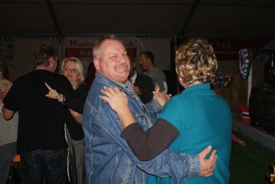 Eine Gruppe von Menschen tanzt in einem Zelt bei einer gesellschaftlichen Veranstaltung, wobei ein Mann und eine Frau im Vordergrund lächelnd und tanzend zu sehen sind.