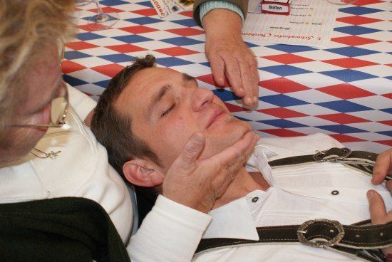 Ein Mann in einem weißen Hemd und einer Hosenträgerhose liegt mit geschlossenen Augen auf einem Tisch, während eine andere Person sein Kinn hält. Eine dritte Person, die eine Brille und eine Halskette trägt, ist in der Nähe. Der Tisch ist mit einer rot, weiß und blau karierten Tischdecke bedeckt.