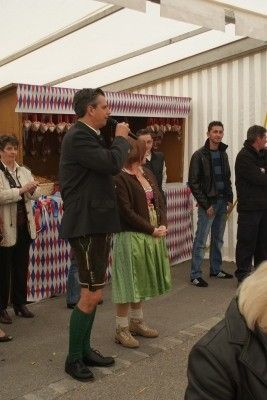 Ein Mann in traditioneller bayerischer Kleidung macht ein Foto mit einer Kamera, während eine Frau in einem grünen Rock neben ihm steht. Sie stehen vor einem Imbissstand mit einem rot-weiß karierten Vordach.