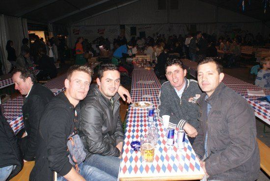Vier Männer sitzen an einem Tisch mit einer karierten Tischdecke, lächeln und posieren für ein Foto. Der Tisch ist mit Getränken und Essen gedeckt, und es sitzen andere Leute an Tischen hinter ihnen.