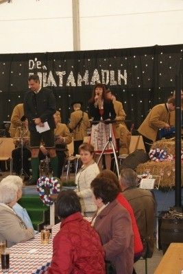 Eine Gruppe von Menschen tritt auf der Bühne mit einem Hintergrund auf, der 'Die Schatzamadin' besagt. Die Darsteller umfassen einen Mann und eine Frau, die singen, mit anderen, die Instrumente spielen. Das Publikum sitzt vor der Bühne.