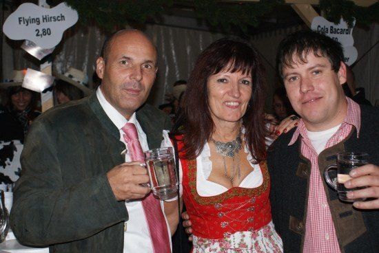 Drei Personen stehen bei einer Veranstaltung zusammen und halten Gläser mit Wasser in der Hand. Der Mann links trägt Anzug und Krawatte, die Frau in der Mitte ein traditionelles Kleid und der Mann rechts ein kariertes Hemd. Hinter ihnen steht ein Schild mit der Aufschrift 'Flying Hirsch 2,80'.