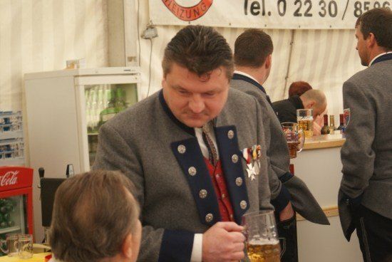 Ein Mann in traditioneller Uniform hält ein Glas Bier in einer Bar-Umgebung, mit anderen Gästen und einem Coca-Cola-Kühler im Hintergrund.