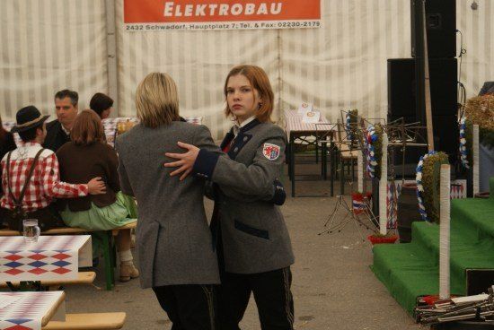 Zwei Frauen in Schuluniformen tanzen zusammen in einem Zelt-Event im Freien. Hinter ihnen sitzen Leute an Tischen, und ein Schild sagt Elektrobau.