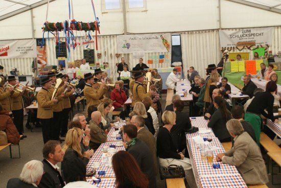 Eine Blaskapelle spielt in einem Zelt-Event mit Tischen, die mit rot-weiß-blau karierten Tischdecken bedeckt sind, während die Leute sitzen und essen.