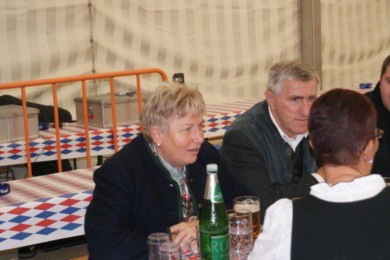 Vier Personen sitzen an einem Tisch in einem Zelt, wobei sich zwei Frauen gegenübersitzen und zwei Männer neben ihnen. Der Tisch ist mit Gläsern, einer Flasche und einer rot-weiß-blauen Tischdecke gedeckt.