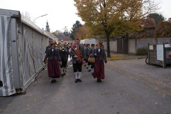 Eine Gruppe von Menschen in traditionellen Kostümen marschiert die Straße entlang, trägt Musikinstrumente und eine Fahne.
