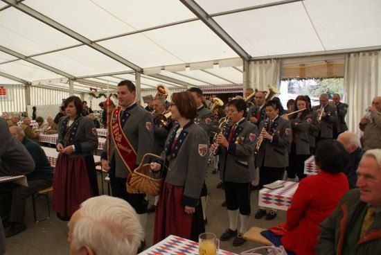 Ein Blasorchester in traditionellen Uniformen spielt in einem Zelt, umgeben von einem Publikum. Die Musiker spielen verschiedene Instrumente, und das Publikum hört aufmerksam zu.