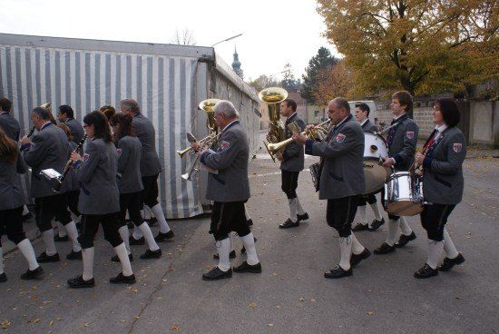 Eine Blaskapelle in traditionellen Uniformen spielt Instrumente im Freien, mit einer gestreiften Wand und Bäumen im Hintergrund.