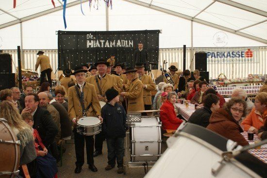 Eine Gruppe von Musikern in traditioneller Kleidung spielt auf Trommeln und anderen Instrumenten in einem großen Zelt, mit einem Publikum, das vor ihnen an Tischen sitzt.