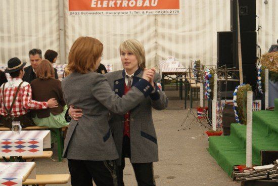 Zwei Frauen tanzen bei einer Outdoor-Veranstaltung zusammen, umgeben von Tischen und Stühlen, mit einer Bühne und Lautsprechern im Hintergrund.