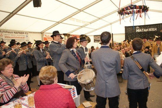 Eine Blaskapelle spielt in einem Zelt, während Zuschauer klatschen, darunter eine Frau in einer roten Jacke. Die Bandmitglieder tragen Uniformen und spielen Instrumente wie Trompeten, Flöten und Trommeln.